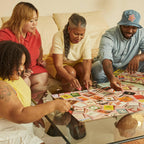 Four friends assemble Match Made in Heaven 1000 Piece Puzzle featuring vintage matchbook covers on a glass table.